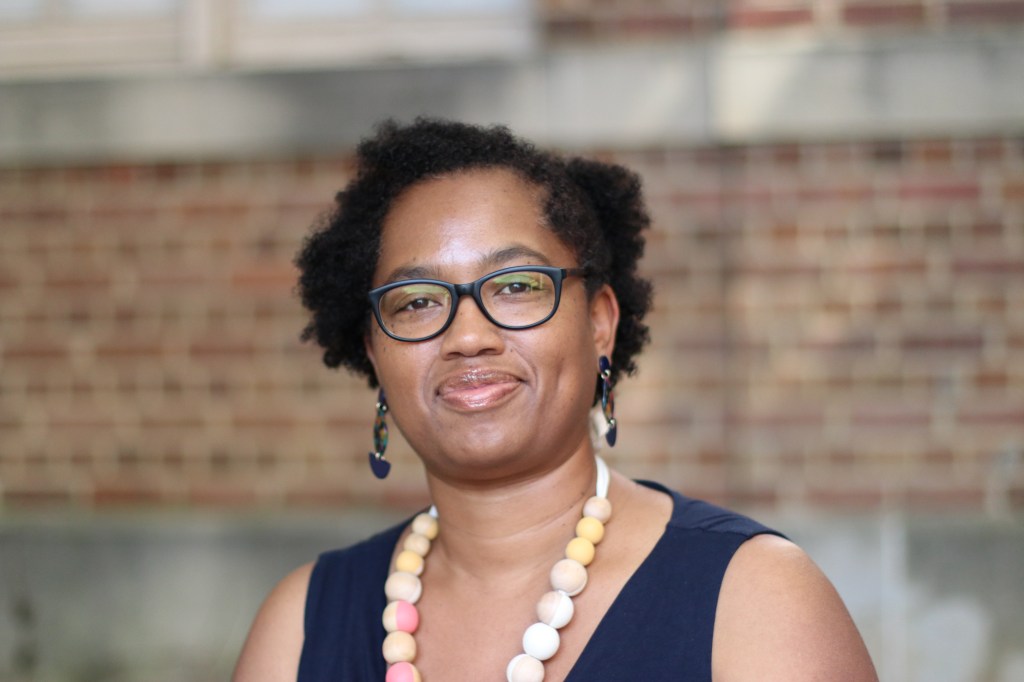 Candace smiles, standing in front of a brown-red brick wall. She is wearing chunky dark brown earrings, black glasses, a wooden bead necklace with pink and white accents, as well as a navy sleeveless dress. 