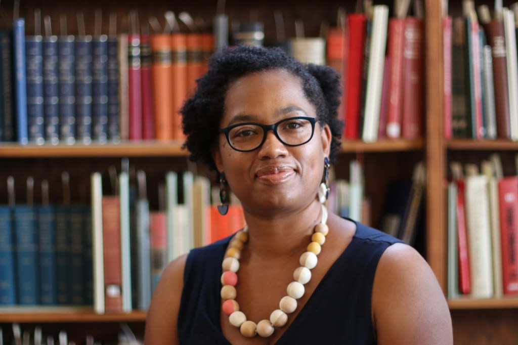Alt-text: Candace smiles, sitting in front of a full bookcase. She is wearing chunky dark brown earrings, black glasses, a wooden bead necklace with pink and white accents, as well as a navy sleeveless dress. 