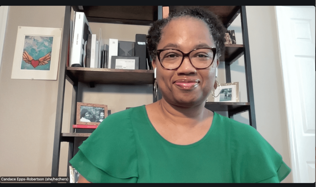 Alt-text: Candace sits in front of a black bookcase with photos and books lining its shelves. She smiles at the camera wearing black glasses and a green shirt with ruffled sleeves. 
