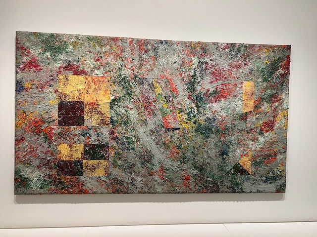 A large abstract painting on a gallery wall. The piece has bold splashes of color and layered textures of paint.